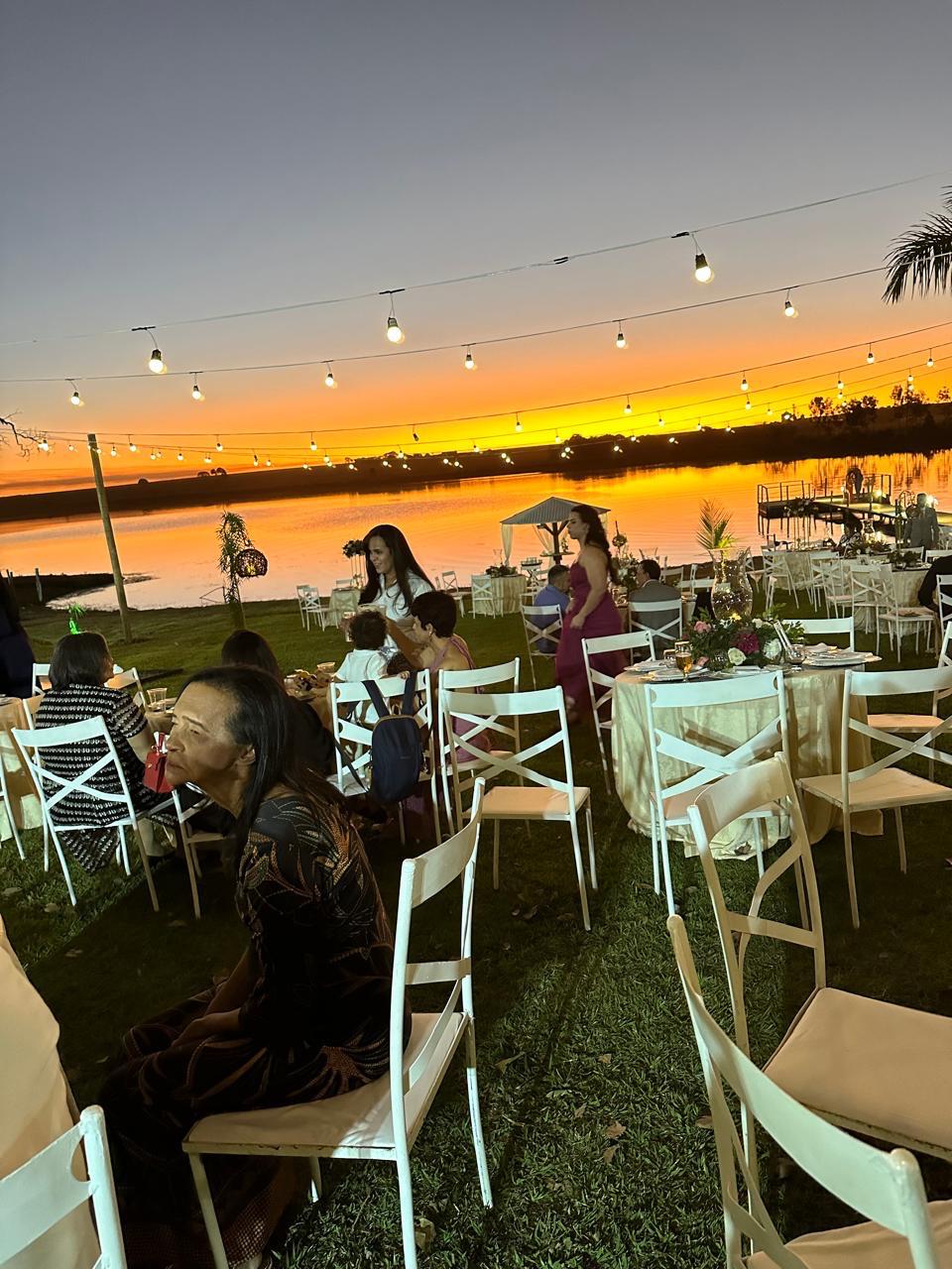 Jantar de recepção noturna ao ar livre com mesas decoradas sob varal de luzes de gambiarra à beira da represa